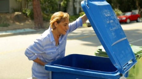 A woman in a blue and white striped shirt stands outside on a sunny day, smiling as she lifts the lid of a large blue recycling bin, possibly awaiting a trash can cleaning service. Other bins and parked cars line the street in the background. A woman in a blue and white striped shirt stands outside on a sunny day, smiling as she lifts the lid of a large blue recycling bin, possibly awaiting a trash can cleaning service. Other bins and parked cars line the street in the background.